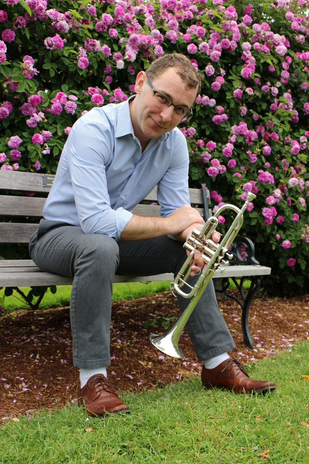 Dylan Prothro seated on a garden bench holding his trumpet, with pink climbing roses behind him