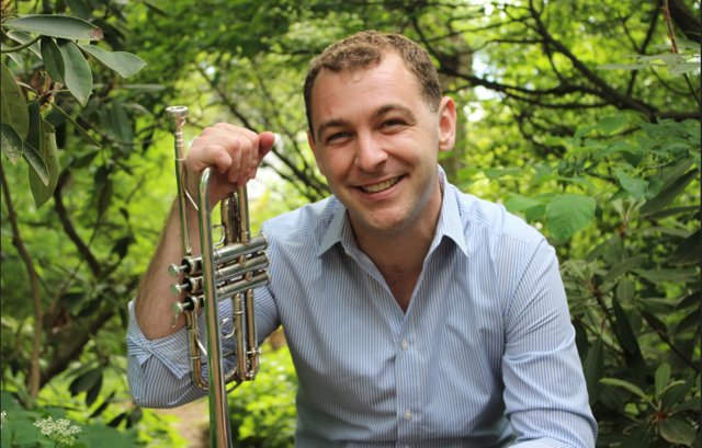 Dylan Prothro smiling, holding his trumpet, against a backdrop of green foliage
